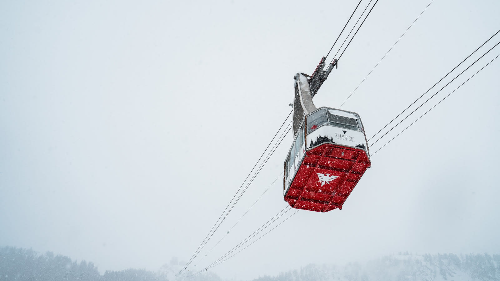Une vue impressionnant en contre bas d'une cabine du téléphérique du Fornet. Le dessous rouge de la cabine avec son logo blanc ressort dans le paysage blanc de l'hiver.